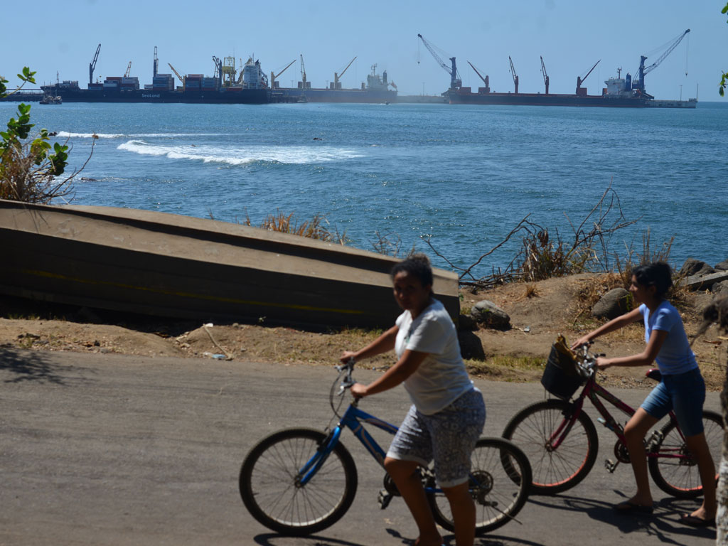 Dos mujeres en bicicleta transitando por una calle costera cerca de una barca, con buques de carga al fondo en el mar.