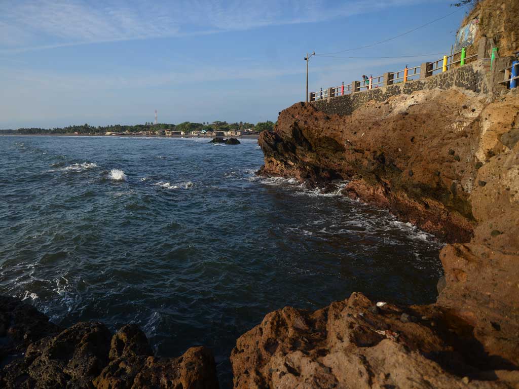 Vista lateral de los acantilados de roca volcánica en Acajutla con el mar rompiendo en la base y el malecón en la parte superior.