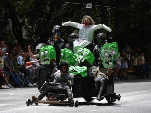 Competidores a bordo de carritos caseros sin motor bajan a toda velocidad por una calle empinada rodeada de público.