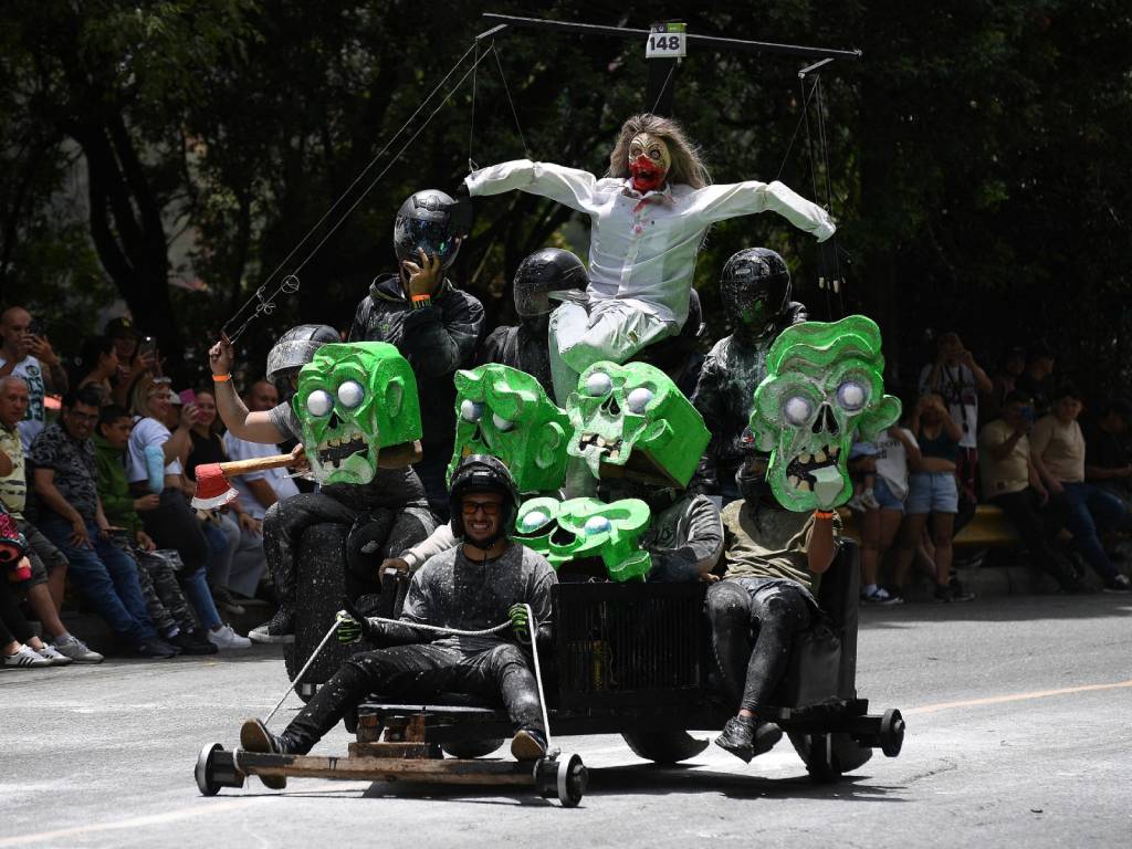 Competidores a bordo de carritos caseros sin motor bajan a toda velocidad por una calle empinada rodeada de público.