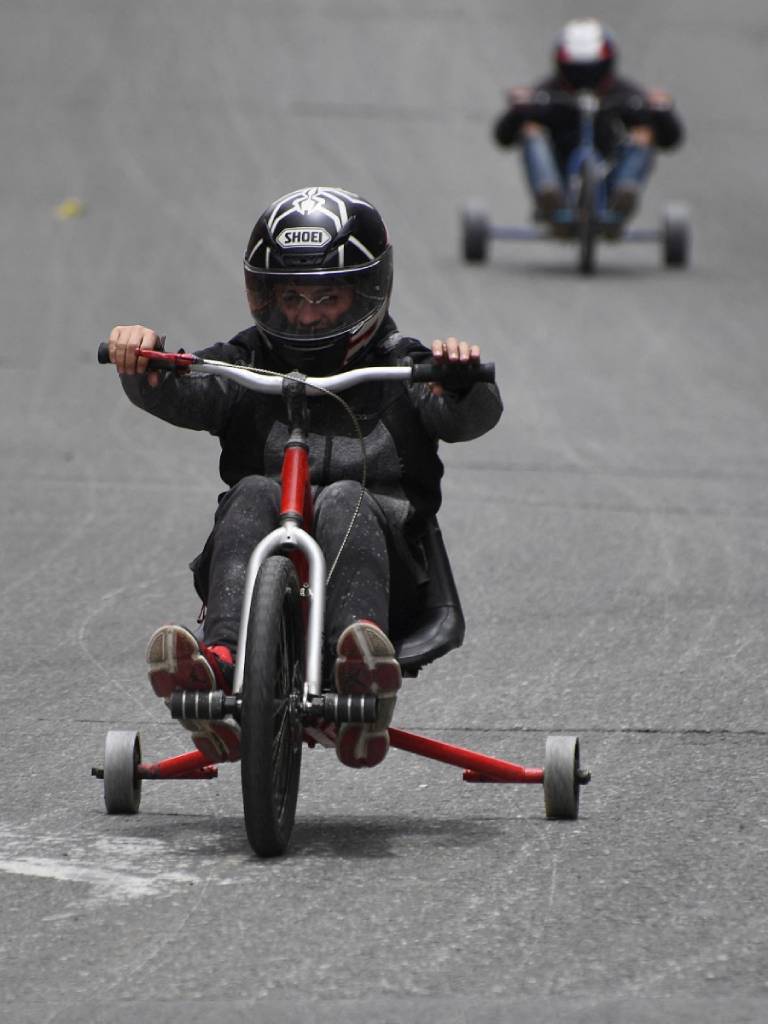 Primer plano de un competidor con casco negro en un Drift Trike rojo y plateado, derrapando en una pista de asfalto, con otro piloto al fondo.