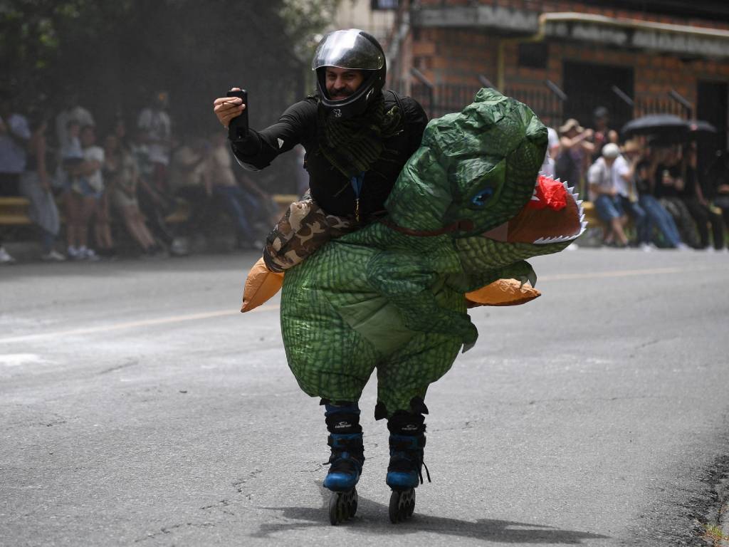 Un patinador disfrazado de dinosaurio verde se toma una foto con su celular mientras avanza por la pista del evento.