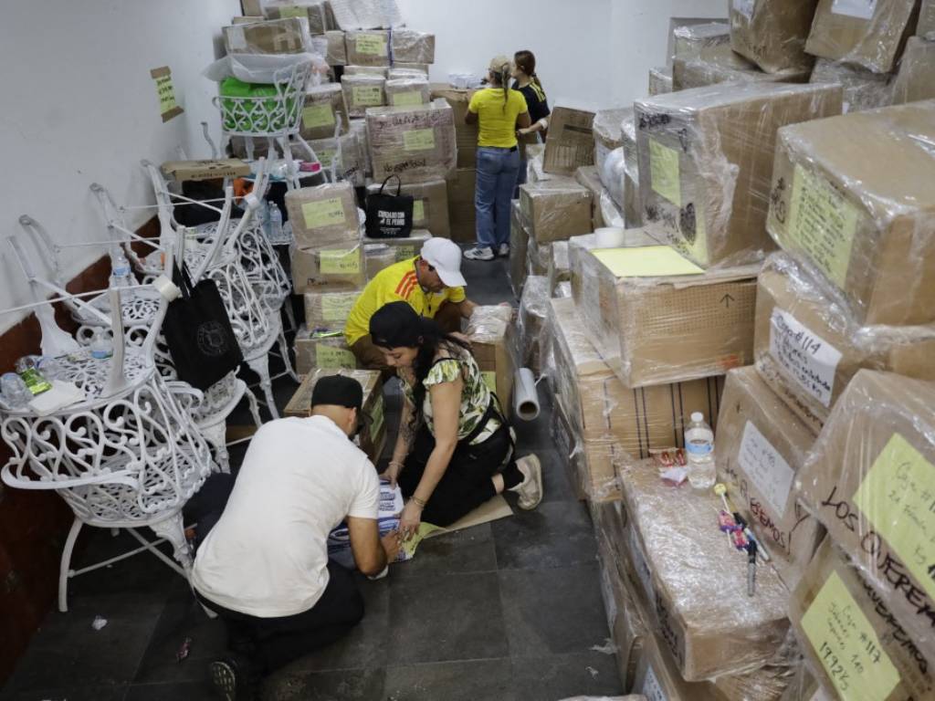 Voluntarios trabajando entre filas de cajas de cartón apiladas y selladas dentro de un centro de acopio.