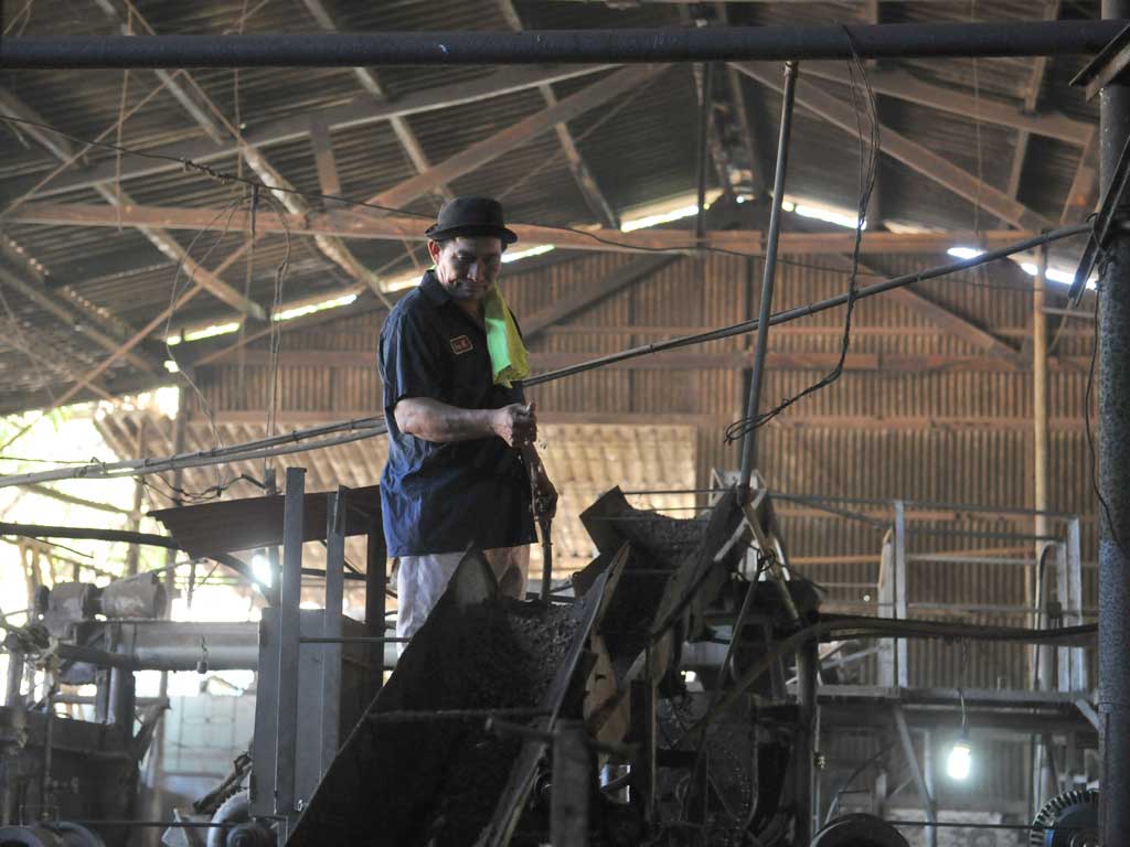 Un hombre con sombrero y toalla en el cuello supervisa la maquinaria industrial desde lo alto, con la estructura metálica de la fábrica de fondo bajo el techo de lámina.