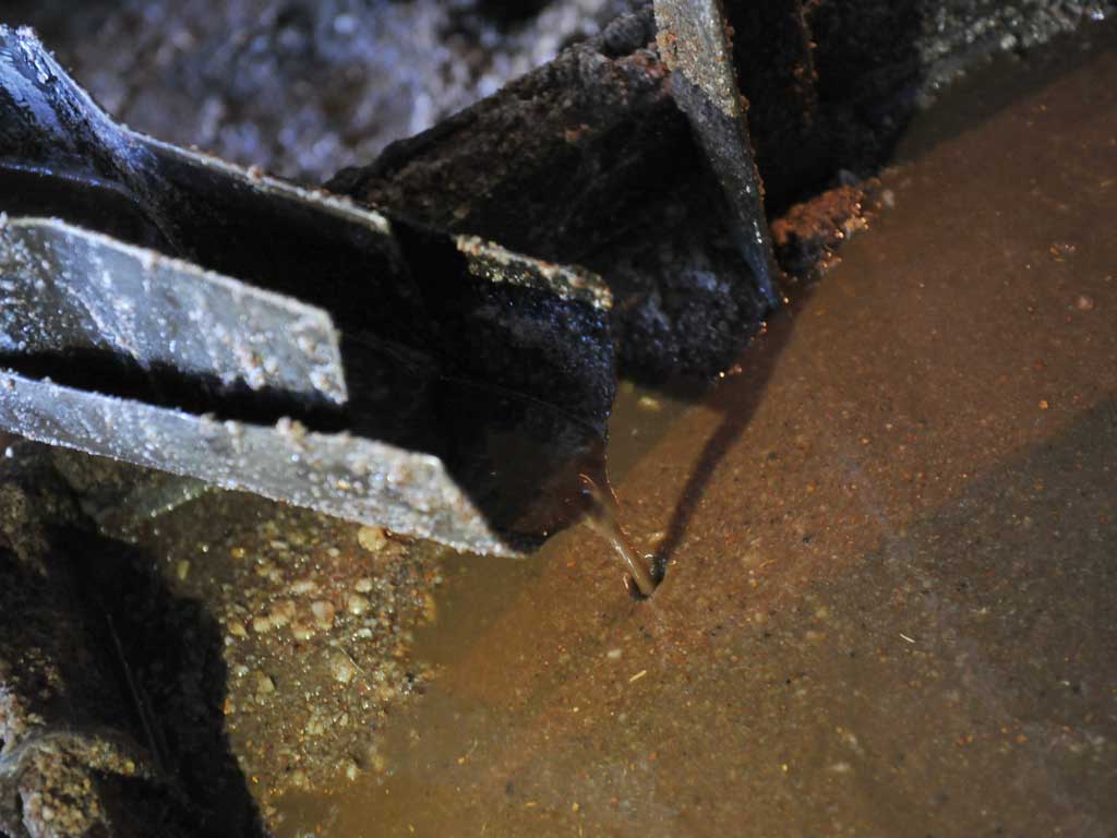 Primer plano de una prensa de metal oxidado donde un pequeño chorro de aceite de coco líquido y turbio cae continuamente desde un canal hacia un recipiente de recolección en el suelo de la fábrica.