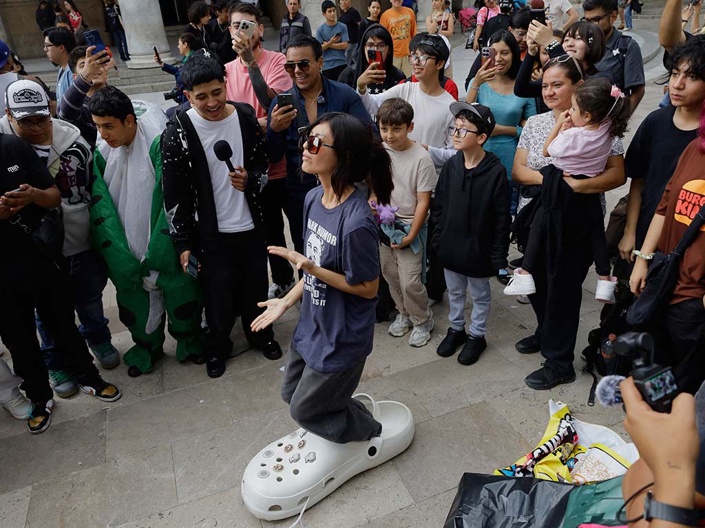 Una joven con gafas oscuras permanece erguida sobre una gran plataforma con forma de Croc blanco mientras el público la graba con celulares.