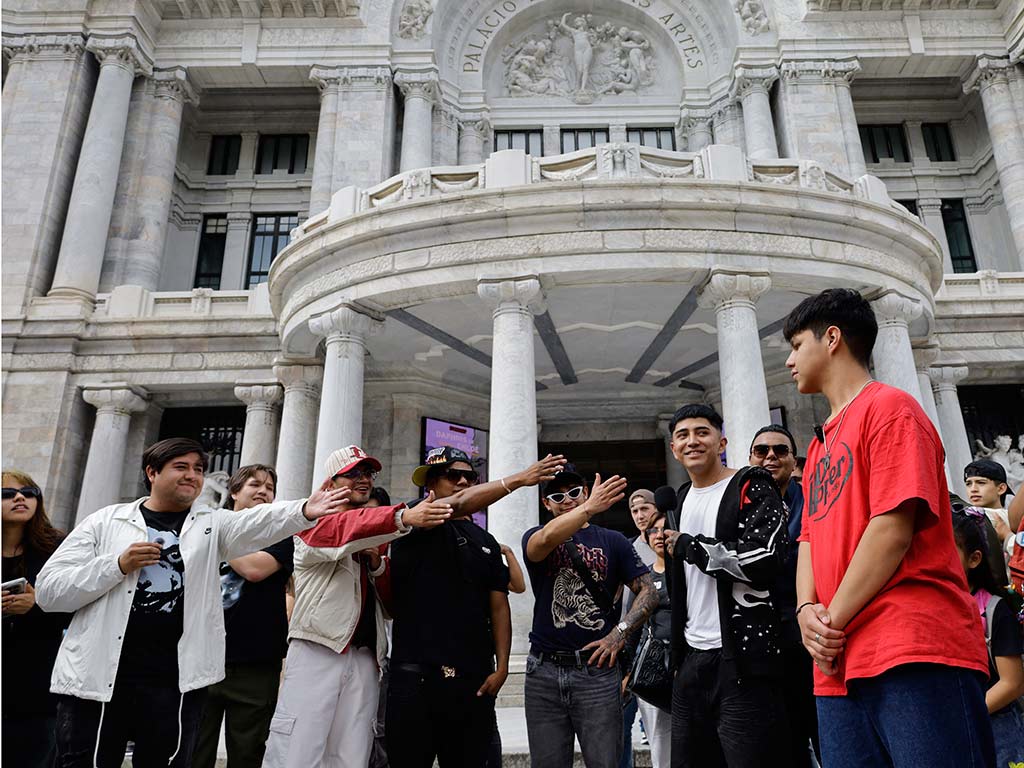 Varios espectadores frente al Palacio de Bellas Artes apuntan con sus brazos extendidos hacia un joven con camiseta roja que permanece parado a la derecha.