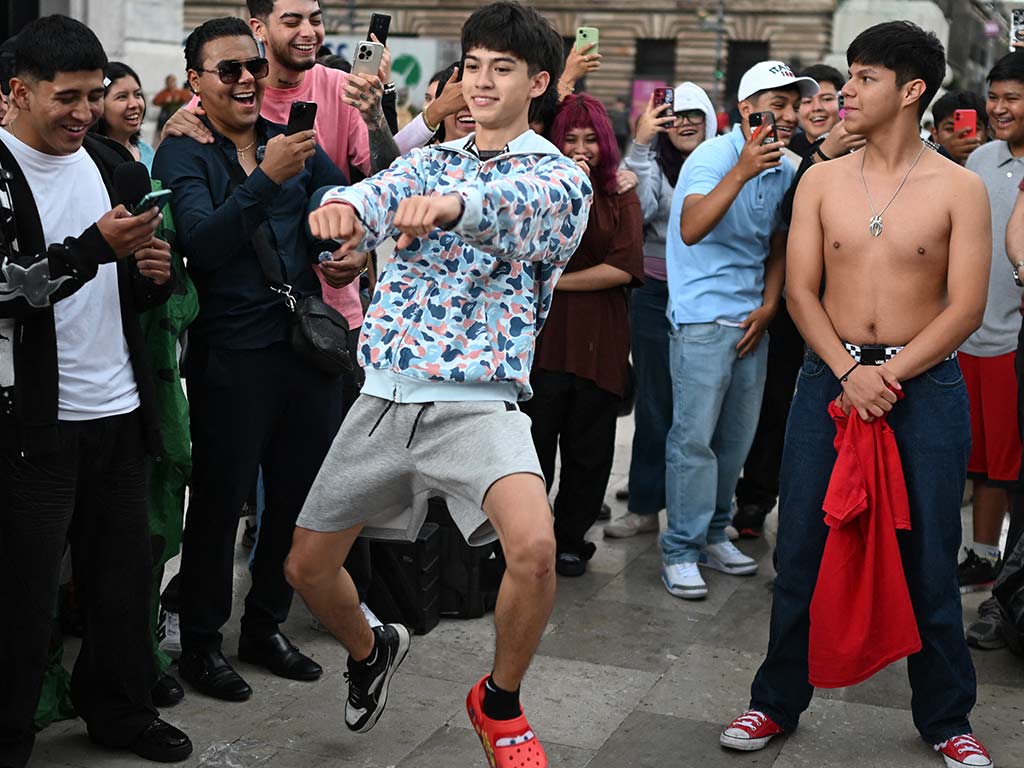 Un joven con sudadera de camuflaje azul y bermudas grises salta sobre un pie en la plaza mientras otro competidor sin camisa lo observa de pie.