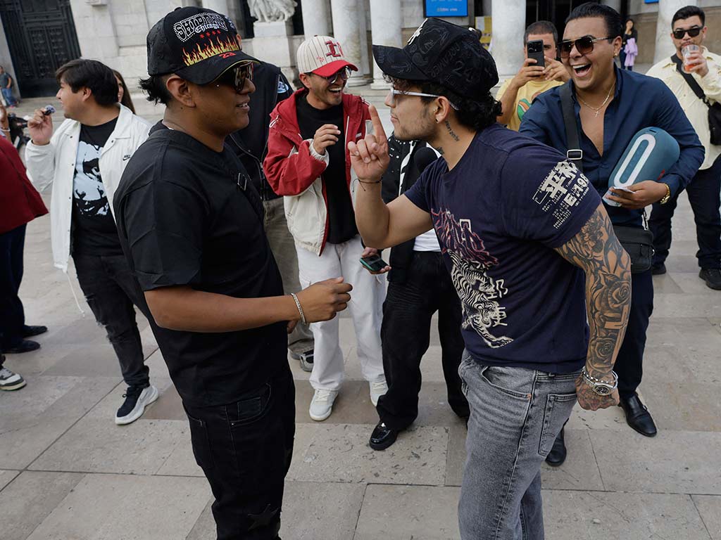 Un joven con camiseta azul con estampados y tatuajes en el brazo levanta el índice señalando hacia el rostro de su rival con gorra negra durante la batalla.