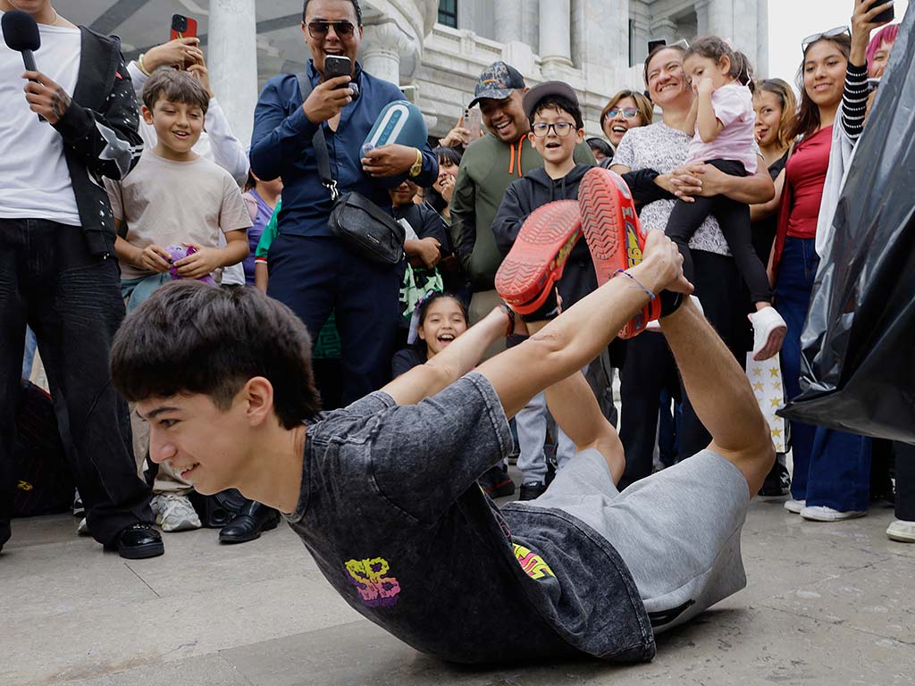 Un joven acostado boca abajo realiza un arco hacia atrás levantando el pecho y las piernas mientras sostiene con sus manos un par de zuecos rojos.