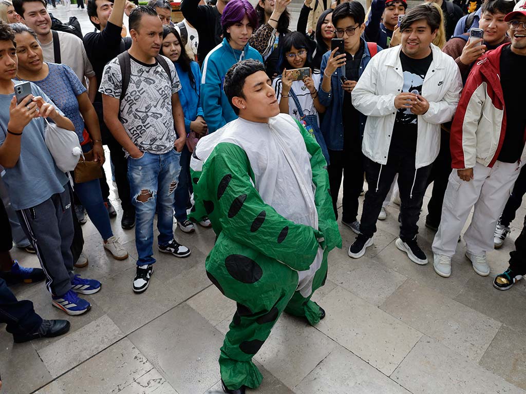 Un joven con disfraz inflable de dinosaurio verde baila en el centro de un círculo de espectadores frente al Palacio de Bellas Artes.