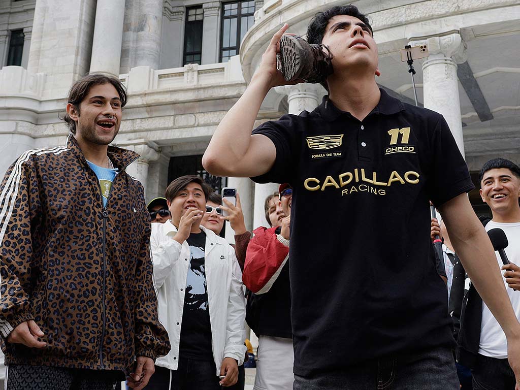 Un joven con polo negra de carreras sostiene un tenis sobre su oreja como si fuera un teléfono, mientras la multitud detrás sonríe.