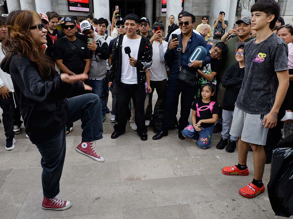 Una joven vestida de negro y tenis rojos se equilibra sobre una pierna levantando la otra en una postura de baile frente a un joven con bermudas.