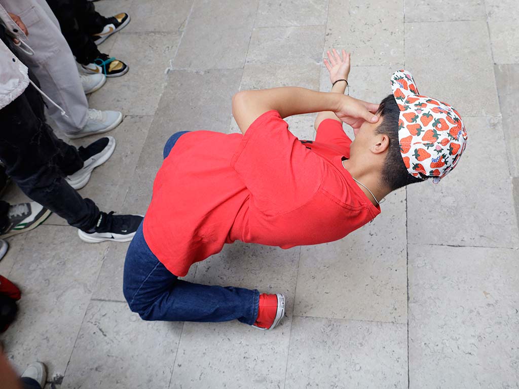 Vista superior de un joven con camiseta roja y gorra estampada de fresas inclinado hacia atrás desde las rodillas sobre el suelo de piedra.
