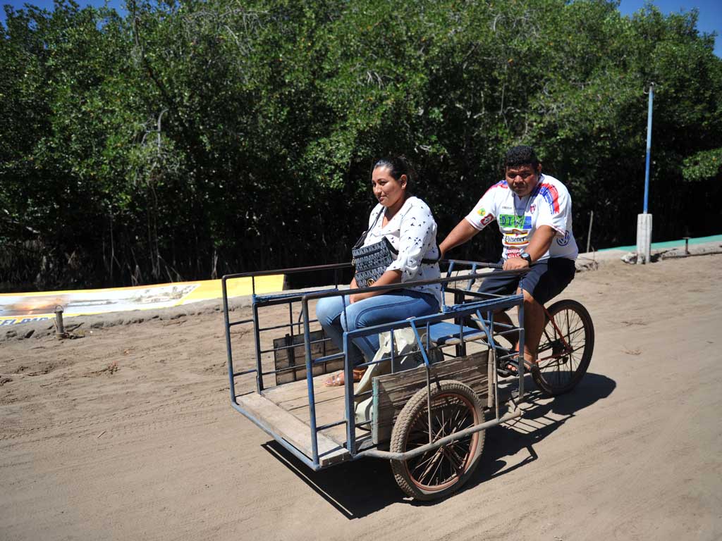 Hombre manejando un triciclo de carga adaptado llevando a una mujer como pasajera por un camino de tierra.