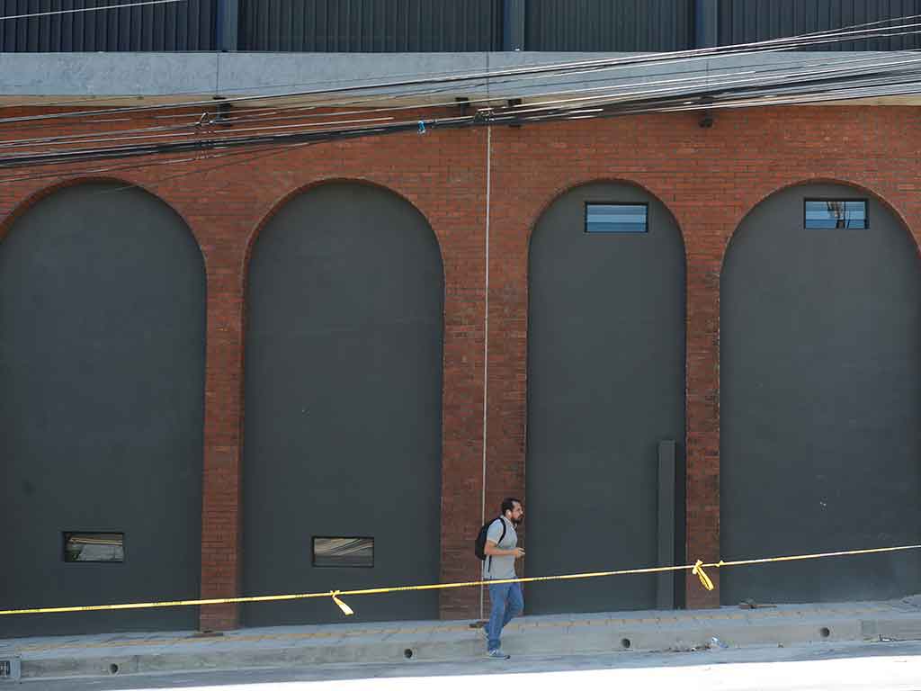 Fotografía que muestra una sección de la fachada del mercado con una pared de ladrillo rojo y una serie de arcos rellenos de un panel gris oscuro. Un peatón con una mochila camina por la acera de delante, detrás de una cinta de precaución amarilla. Se ven cables eléctricos en la parte superior.