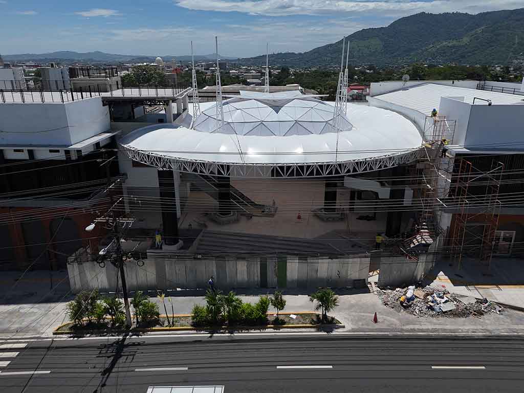 Vista frontal de ángulo medio de la fachada del Mercado Cuscatlán, destacando la gran estructura de techo blanco cónico y las columnas negras. Hay andamios en el lado derecho y la calle 25 Avenida Sur en primer plano.