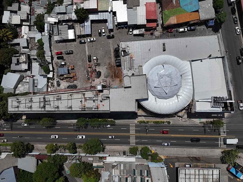 Fotografía aérea a vista de pájaro que muestra el complejo del Mercado Cuscatlán y sus alrededores. Se distingue la estructura de techo blanco y circular, las áreas de estacionamiento adyacentes, las calles con tráfico y las zonas residenciales y comerciales circundantes. Los andamios son visibles en varias partes del edificio.