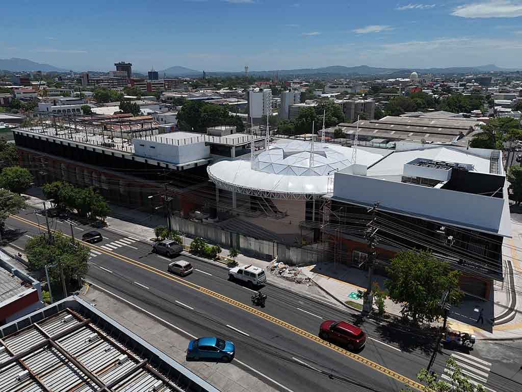 Vista aérea de ángulo medio del Mercado Cuscatlán en remodelación, mostrando el techo blanco cónico central y las estructuras laterales modernizadas. Se ve el tráfico en la 25 Avenida Sur y el horizonte de la ciudad de San Salvador bajo un cielo azul con nubes.