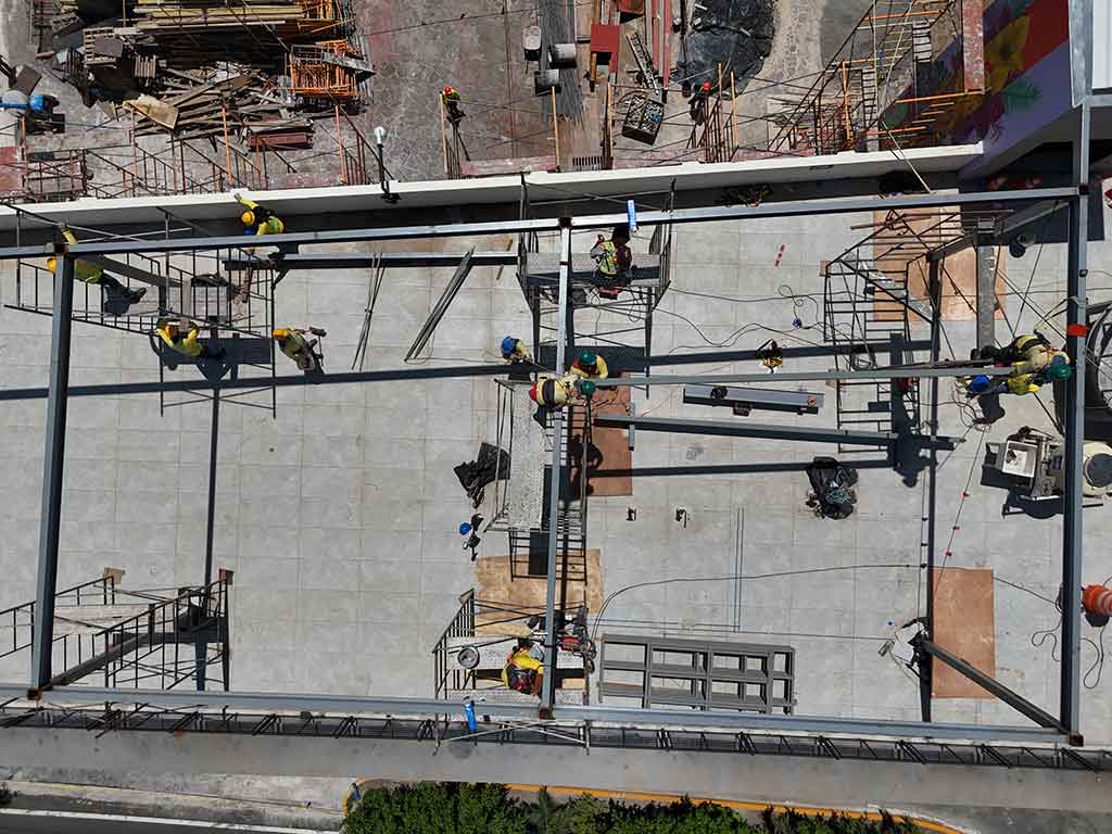 Vista aérea de ángulo alto de trabajadores de la construcción con cascos amarillos montando una estructura de vigas de acero sobre el techo del Mercado Cuscatlán. Abajo se ve el pavimento de adoquines y edificios circundantes.