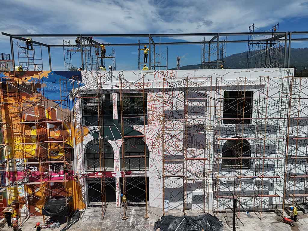 Vista lateral de ángulo medio de la fachada del mercado, cubierta por una red de andamios. A la izquierda, se empieza a ver un mural colorido con rostros. Trabajadores se mueven por los andamios y en la parte superior.