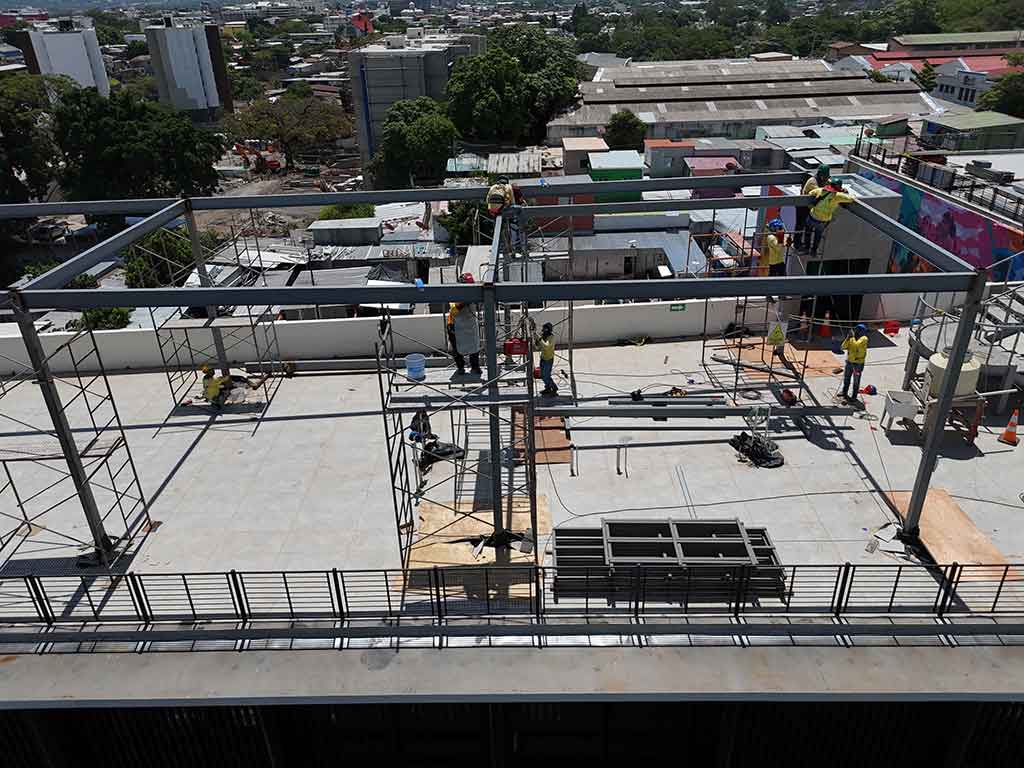 Fotografía de ángulo medio que muestra la fachada principal en remodelación del Mercado Cuscatlán, con su distintiva estructura de techo blanco y columnas negras. Hay andamios en el lateral derecho y cercas de lámina galvanizada en la acera. Un trabajador con casco azul pasa por delante.