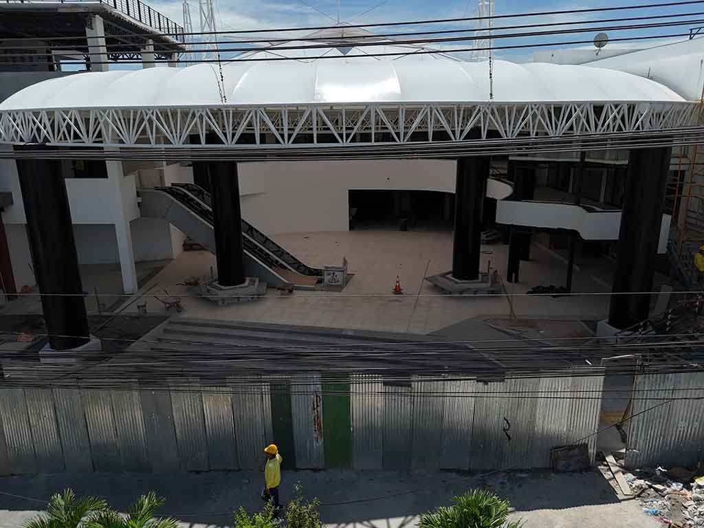 Vista frontal de ángulo medio de la entrada principal del mercado a nivel de calle. Grandes columnas negras sostienen la estructura del techo. Se ven escaleras mecánicas en el interior. Un trabajador con casco amarillo pasa por delante en primer plano.