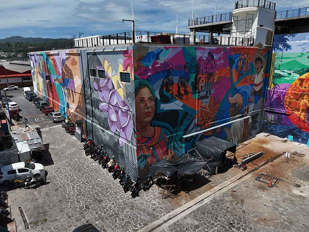 Vista aérea de ángulo medio del mural colorido pintado en la pared lateral del Mercado Cuscatlán. El mural muestra rostros humanos, flores y escenas tradicionales. Hay motocicletas estacionadas abajo y un auto blanco en la calle.