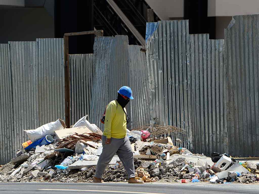 Primer plano de un trabajador de la construcción con camisa amarilla de manga larga, pantalones grises, un pasamontañas negro y un casco azul, caminando de derecha a izquierda por delante de una cerca de lámina galvanizada. Hay una pila de escombros de construcción en primer plano.
