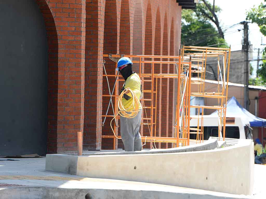 Fotografía que destaca una serie de arcos de ladrillo rojo en la base de la estructura del mercado. Un trabajador con camisa amarilla y casco azul está de pie detrás de los arcos, cerca de unos andamios de color naranja, sosteniendo un rollo de cable amarillo. Una rampa de hormigón curva está en primer plano.