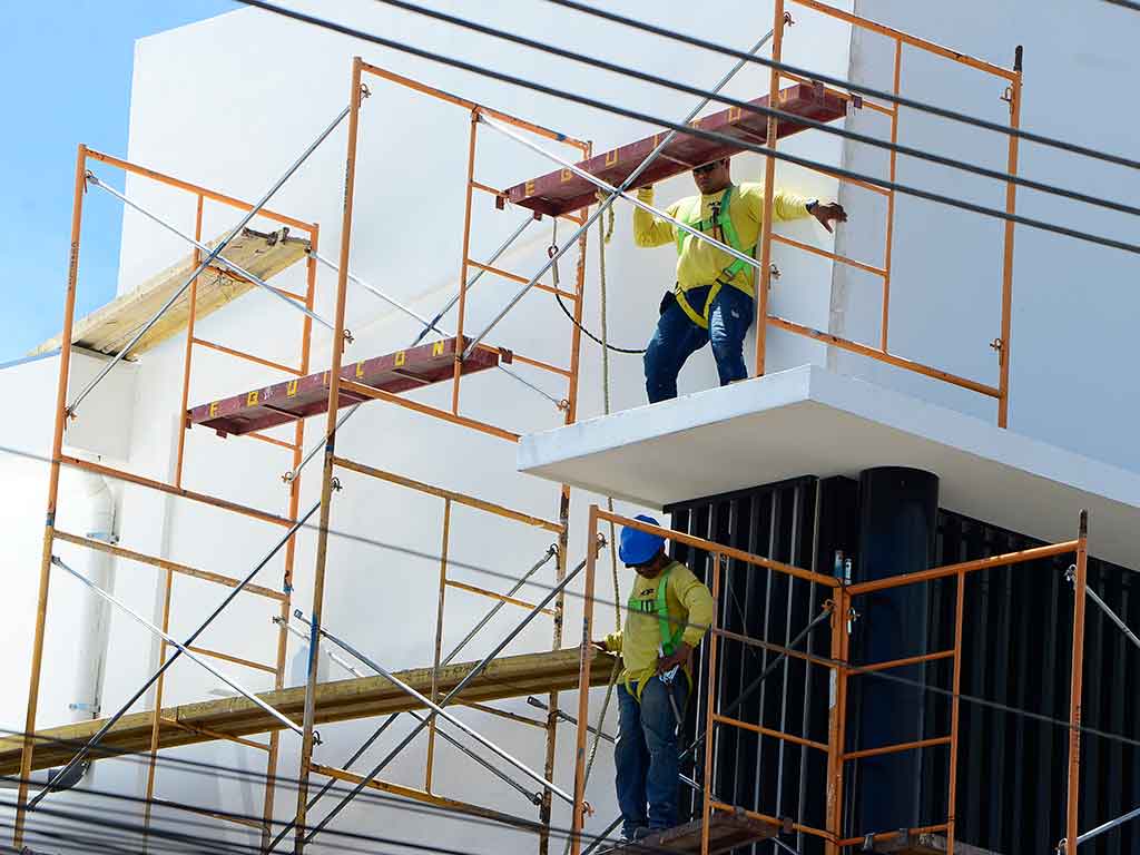 Vista en ángulo hacia arriba de la parte superior del edificio del mercado, mostrando andamios amarillos y naranjas. Dos trabajadores están en los andamios; uno está en una plataforma superior y el otro más abajo. Se ven cables eléctricos y el cielo azul.