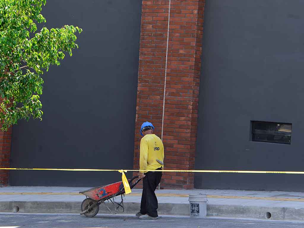 Fotografía de ángulo bajo que muestra a un trabajador de la construcción de espaldas, con camisa amarilla y casco azul, empujando una carretilla roja con materiales a lo largo de un camino delimitado por una cinta de precaución amarilla. El fondo es una pared gris lisa con una columna de ladrillos rojos