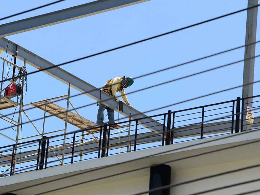Vista en ángulo hacia arriba de un trabajador con equipo de seguridad (arnés, casco verde) soldando una viga de acero en el techo del edificio. Hay andamios a la izquierda y cables eléctricos cruzando la escena contra un cielo azul claro.