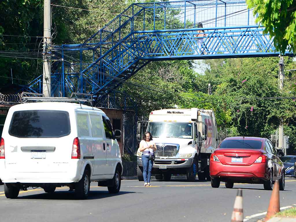 Una mujer con camiseta rosa y jeans está parada en el carril central del Bulevar Tutunichapa, entre un panel blanco y un camión de basura que avanza. Al fondo, una pasarela peatonal azul cruza sobre la vía. La imagen resalta el riesgo que toma el peatón al no usar la pasarela en una zona de tráfico denso en San Salvador.
