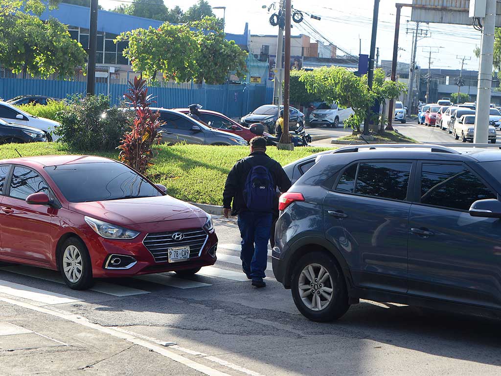 Una fotografía a nivel de calle muestra a un peatón con una mochila azul cruzando un paso de cebra en la concurrida Alameda Juan Pablo II en San Salvador. Camina entre un auto Hyundai sedán rojo a su izquierda y un SUV oscuro a su derecha. Ambos vehículos están detenidos pero listos para avanzar. El fondo muestra la gasolinera Puma Constitución y tráfico adicional.