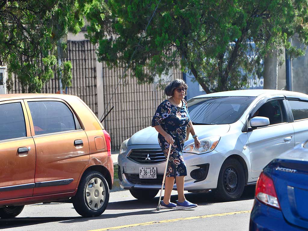 Fotografía de una mujer mayor que viste un vestido floral y usa un bastón para ayudarse a caminar. Está cruzando un paso de cebra en la Avenida Bernal en San Salvador, pasando por delante de un sedán plateado detenido en el tráfico. Un auto naranja y otro azul oscuro también son visibles.