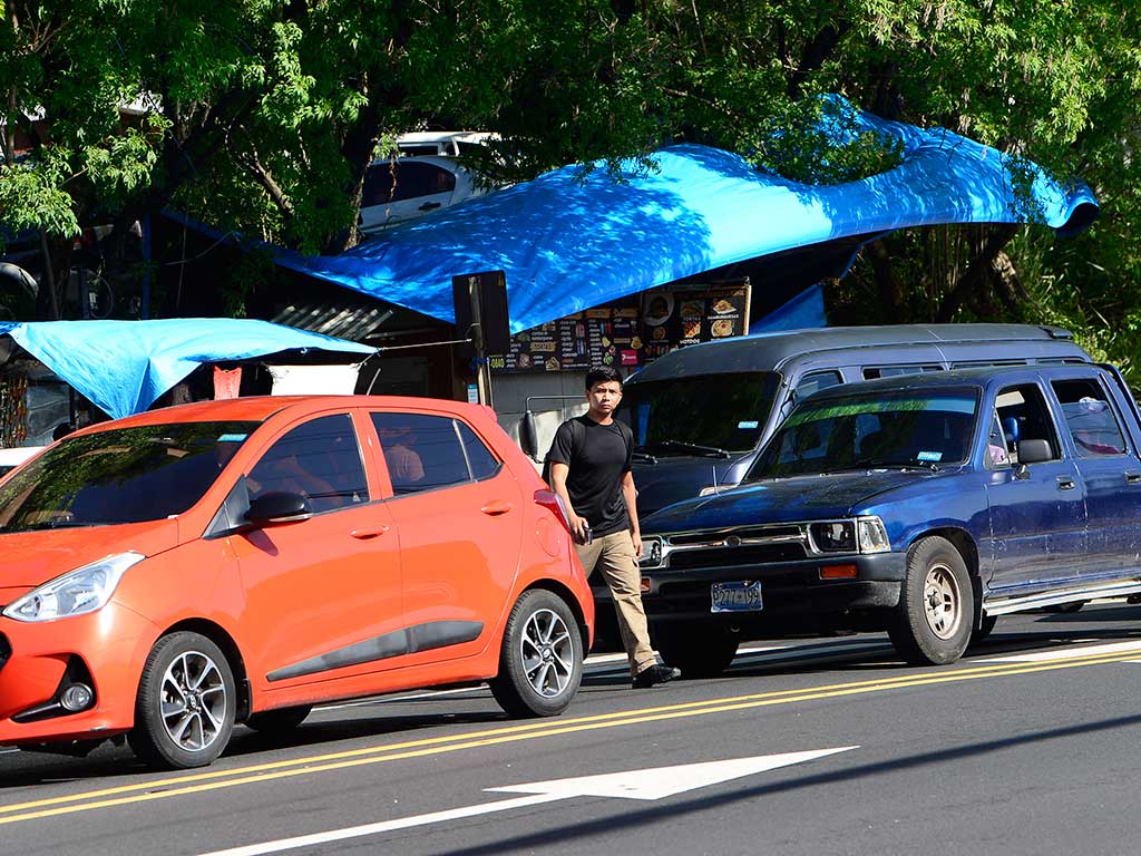 Fotografía que captura a un joven con una camiseta negra y pantalones cortos cruzando una concurrida vía en San Salvador, la Avenida Bernal. Camina entre un auto naranja a su izquierda y una camioneta pick-up azul a su derecha. Ambas están detenidas en el tráfico. Al fondo, se ven árboles frondosos y una estructura con un techo azul.