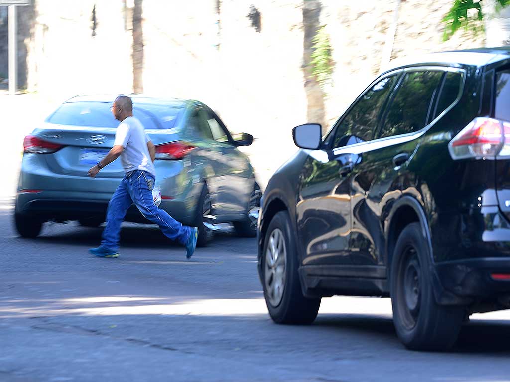 Una toma de acción a nivel de calle muestra a un hombre con camiseta blanca y jeans corriendo para cruzar la Avenida Don Bosco. Un sedán gris pasa detrás de él y un SUV negro está detenido en primer plano a la derecha. La escena evoca movimiento y un cruce riesgoso en San Salvador.