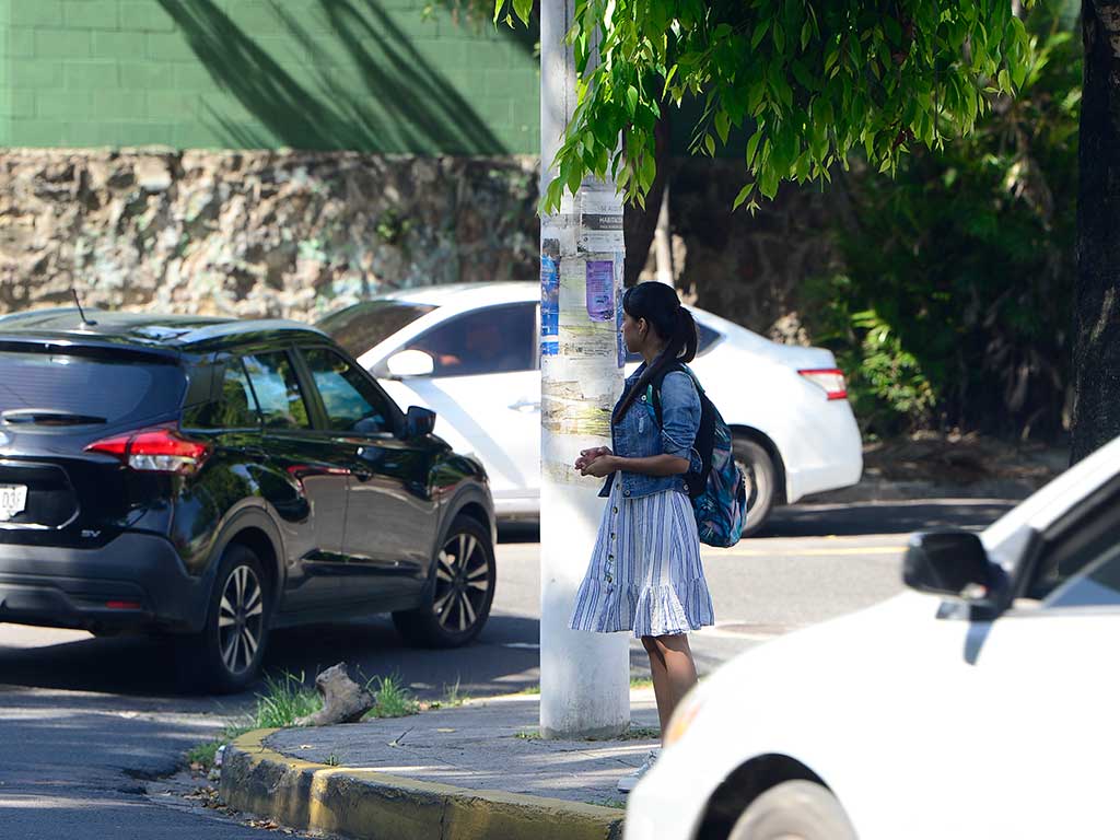Una joven con una falda a rayas, chaqueta de mezclilla y mochila espera en una esquina de la calle cerca de un poste de luz. Está de pie junto al Bulevar Universitario en San Salvador. Al fondo se ven autos detenidos y un muro de piedra con vegetación. La imagen resalta la espera de un peatón en una vía transitada.