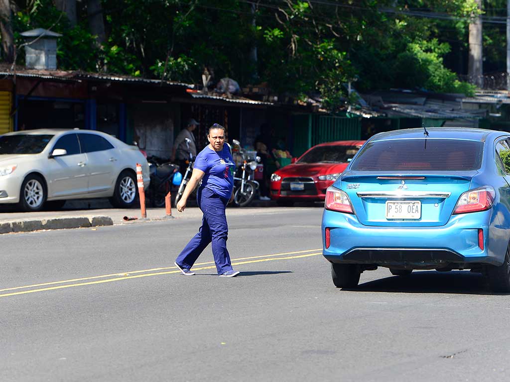 Una mujer con uniforme azul (camiseta y pantalón) cruza el Bulevar Tutunichapa en San Salvador. Camina sobre la doble línea amarilla que divide los carriles. Un sedán azul con placa P 58 0EA está detenido a su derecha y un sedán blanco a su izquierda en la acera. Al fondo hay puestos de venta informales y árboles.