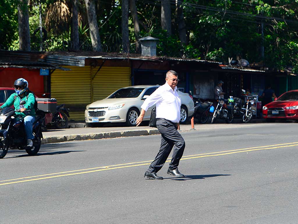 Un hombre con camisa blanca de vestir y pantalones negros cruza el Bulevar Tutunichapa. Está en medio de la calle, entre carriles de tráfico. A la izquierda, un motociclista con casco verde avanza. Al fondo se ven puestos de venta y árboles. La foto captura el movimiento del peatón en una calle concurrida de San Salvador.