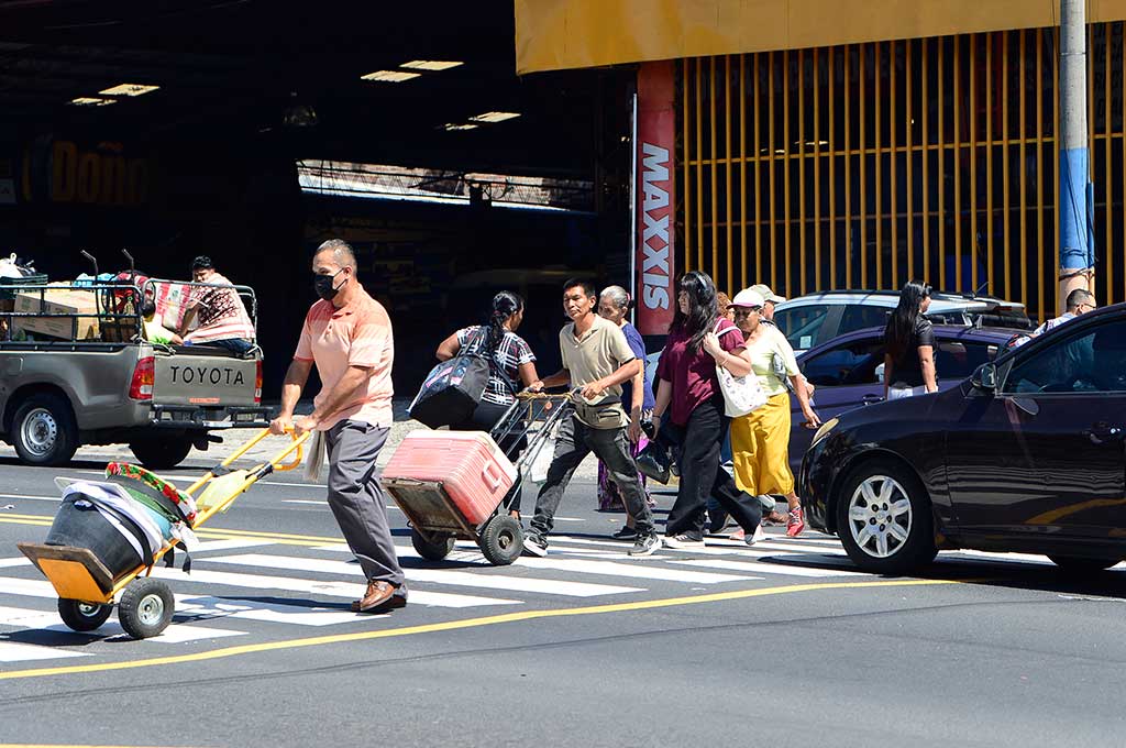 Una escena de calle concurrida en la intersección del Bulevar Venezuela y la Avenida 29 de Agosto, San Salvador. Un hombre empuja una carretilla con mercancía, mientras otros peatones cruzan la calle en diferentes direcciones. Hay una camioneta pick-up con personas en la cama y un auto negro cerca. El fondo muestra un edificio comercial con el letrero "MAXXIS". La imagen evoca el caos del comercio y el tráfico.