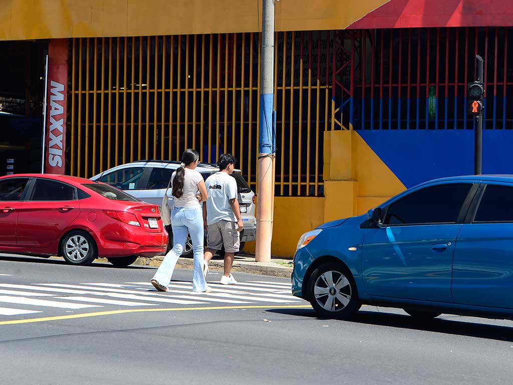 Una pareja de jóvenes cruza el Bulevar Venezuela cerca de la Avenida 29 de Agosto. Caminan por un paso de cebra, pero fuera de las líneas. Un sedán azul avanza hacia la derecha y un auto rojo está estacionado detrás. Al fondo se ve un edificio amarillo con rejas y el letrero "MAXXIS". La foto captura el cruce en una zona de tráfico y paradas de buses en San Salvador.