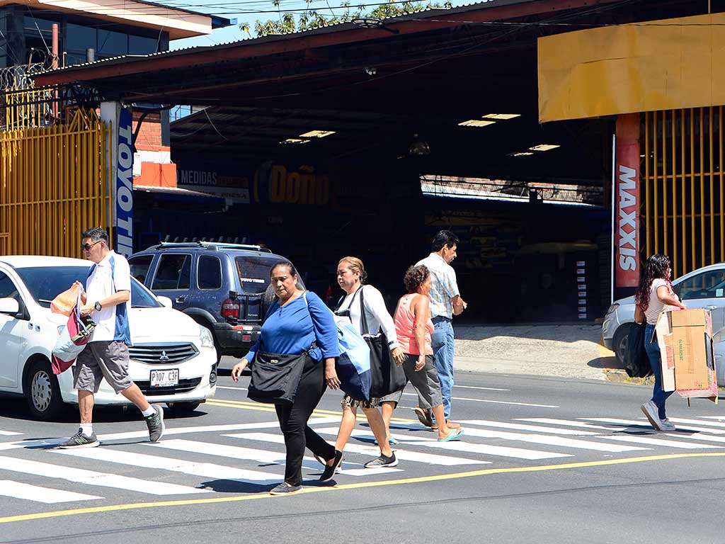 Un grupo de peatones cruza el Bulevar Venezuela en un paso de cebra cerca de la Avenida 29 de Agosto. Varias personas caminan por las líneas blancas, mientras un sedán blanco con placa P 107 C3F está detenido detrás. Al fondo se ven edificios comerciales con letreros como "TOYO TIRES" y "MAXXIS". La imagen muestra el cruce peatonal en una zona transitada y de riesgo en San Salvador.