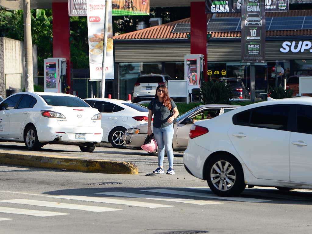 Fotografía de una mujer con jeans, una camisa oscura y zapatos deportivos, parada en un separador central angosto de la Alameda Juan Pablo II. Sostiene un casco rosa de motociclista. Está rodeada por ambos lados de vehículos blancos en movimiento o detenidos en el tráfico, frente a la gasolinera Puma Constitución.