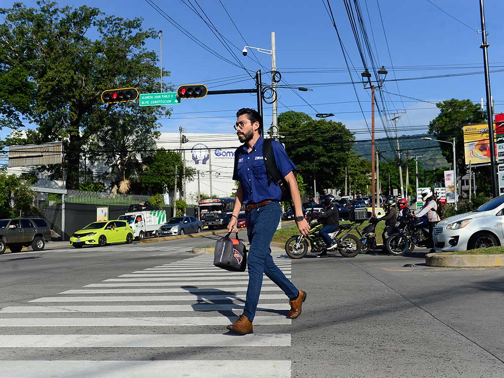 Imagen de un hombre joven con una camisa de vestir azul, jeans y botas marrones cruzando un paso de cebra en la intersección de la Alameda Juan Pablo II con el Bulevar Constitución. Sostiene un bolso. Al fondo se ve tráfico de vehículos, incluyendo autos y motocicletas, detenidos por el semáforo vehicular que muestra luz roja.