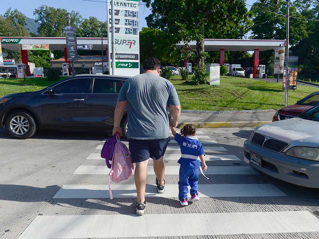 Toma trasera de un hombre con una camiseta gris y pantalones cortos oscuros cruzando un paso de cebra con un niño pequeño que viste un uniforme azul de la escuela o de artes marciales. El hombre lleva una mochila rosa. Cruzan la Alameda Juan Pablo II en San Salvador, con tráfico detenido a su izquierda y frente a ellos la entrada a la gasolinera Puma Constitución.