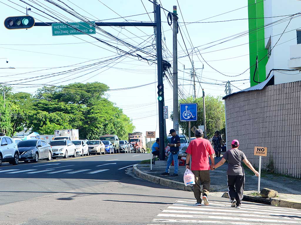 Fotografía de una intersección en San Salvador, con un letrero de calle elevado que dice "AVENIDA BERNAL" y "BOULEVARD UNIVERSITARIO". Dos peatones caminan por el paso de cebra: un hombre con camisa roja y pantalones cortos cruzando primero, seguido de una mujer con camisa gris y pantalones oscuros. Hay tráfico detenido en múltiples carriles y muchos cables eléctricos aéreos.