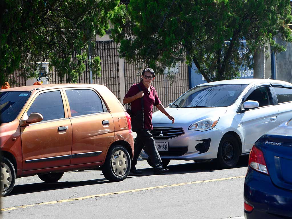 Fotografía que muestra a un hombre con una camiseta granate y pantalones oscuros cruzando un paso de cebra en la Avenida Bernal en San Salvador. Camina entre dos autos detenidos: un hatchback naranja a su izquierda y un sedán plateado a su derecha. El fondo muestra árboles frondosos.