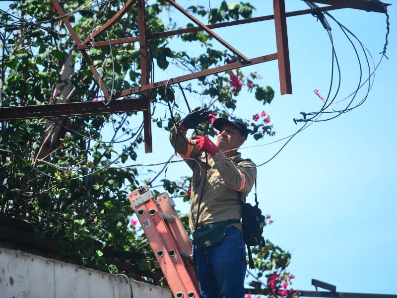 Un trabajador sobre una escalera manipula y corta cables eléctricos colgados de una estructura metálica en la vía pública.